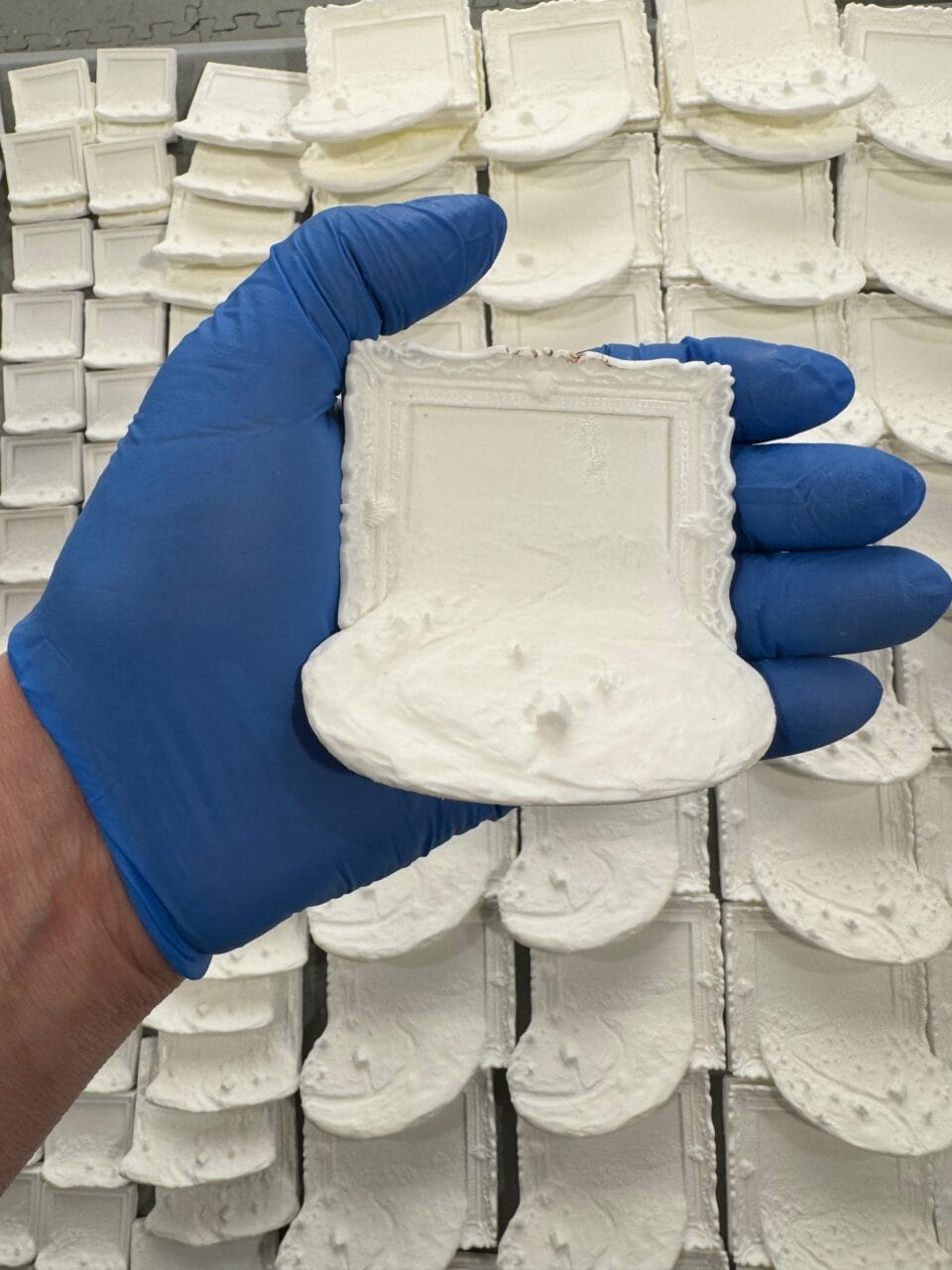 a gloved hands holds up a blank resin cast of the a miniature landscape painting spilling out of an ornate frame. there are dozens more casted in the background, waiting to be painted