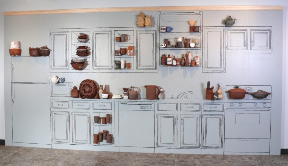a gallery wall outlined with kitchen cabinets and counters, with three-dimensional reliefs of ceramic vessels and objects installed on the wall