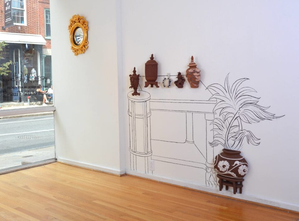 an overview of an installation in a gallery showing a group of ceramic sculptural reliefs of vases, decorative vessels, and other objects on a wall, against a line drawing of a mantle in a gallery space