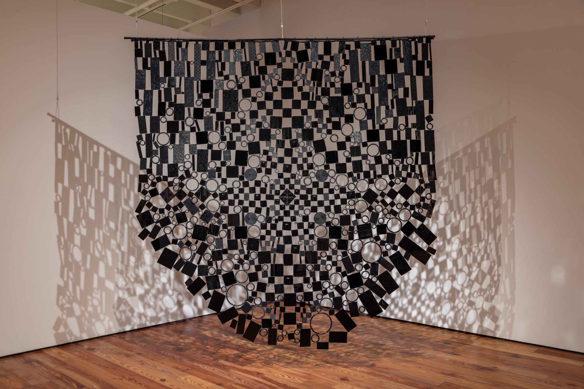 a suspended mixed-media tapestry installation made of black pieces of material, casting shadows on a gallery wall