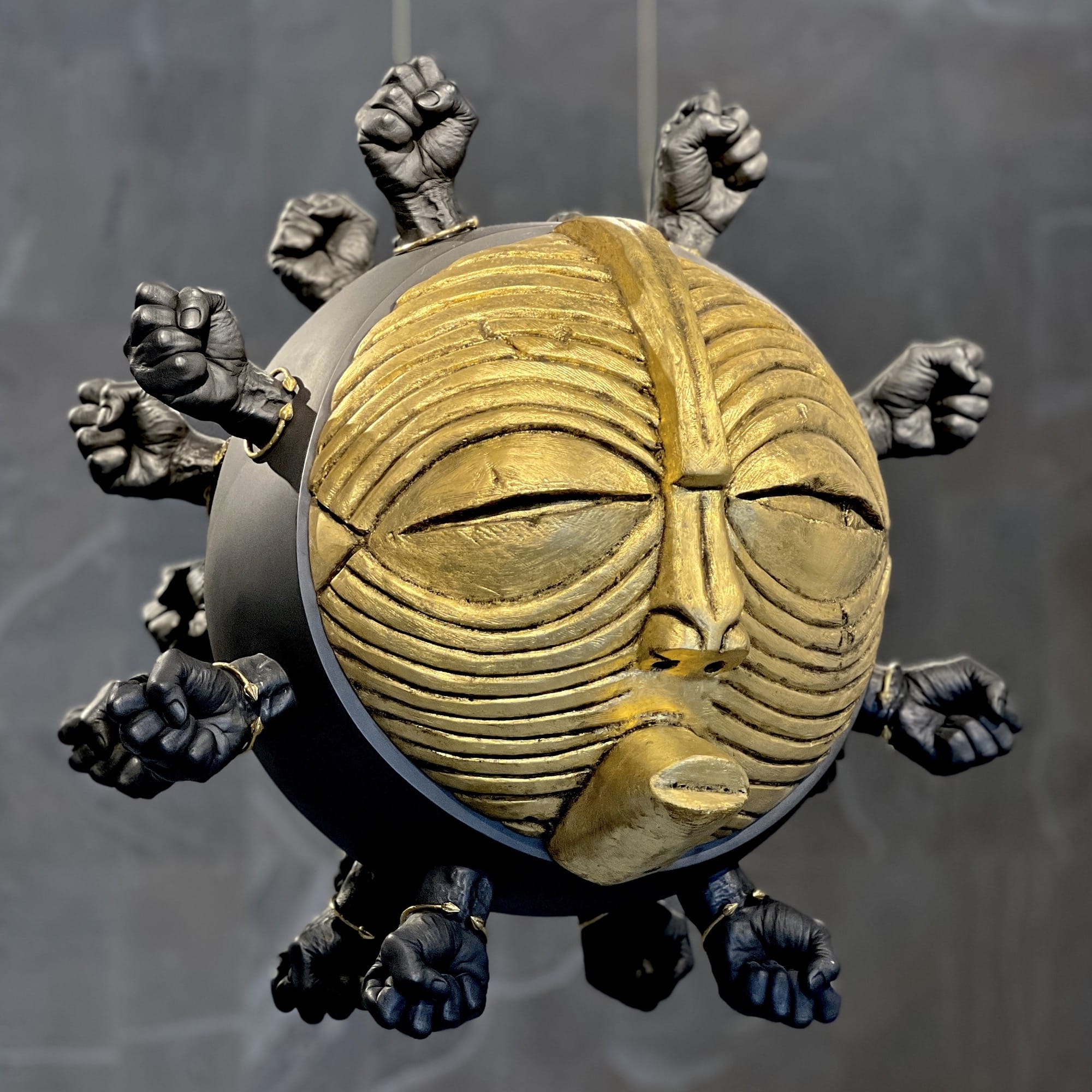 a suspended sculpture of a sphere with an African mask on the front and nodes of power fists, overall resembling a virus