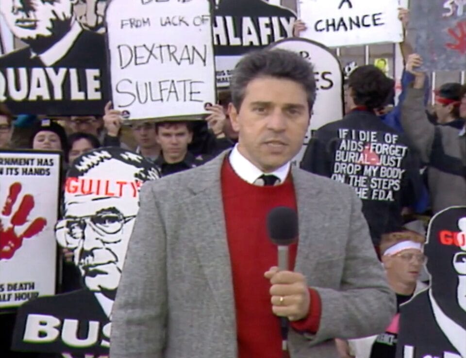 a man stands speaking to the camera in front of a protest