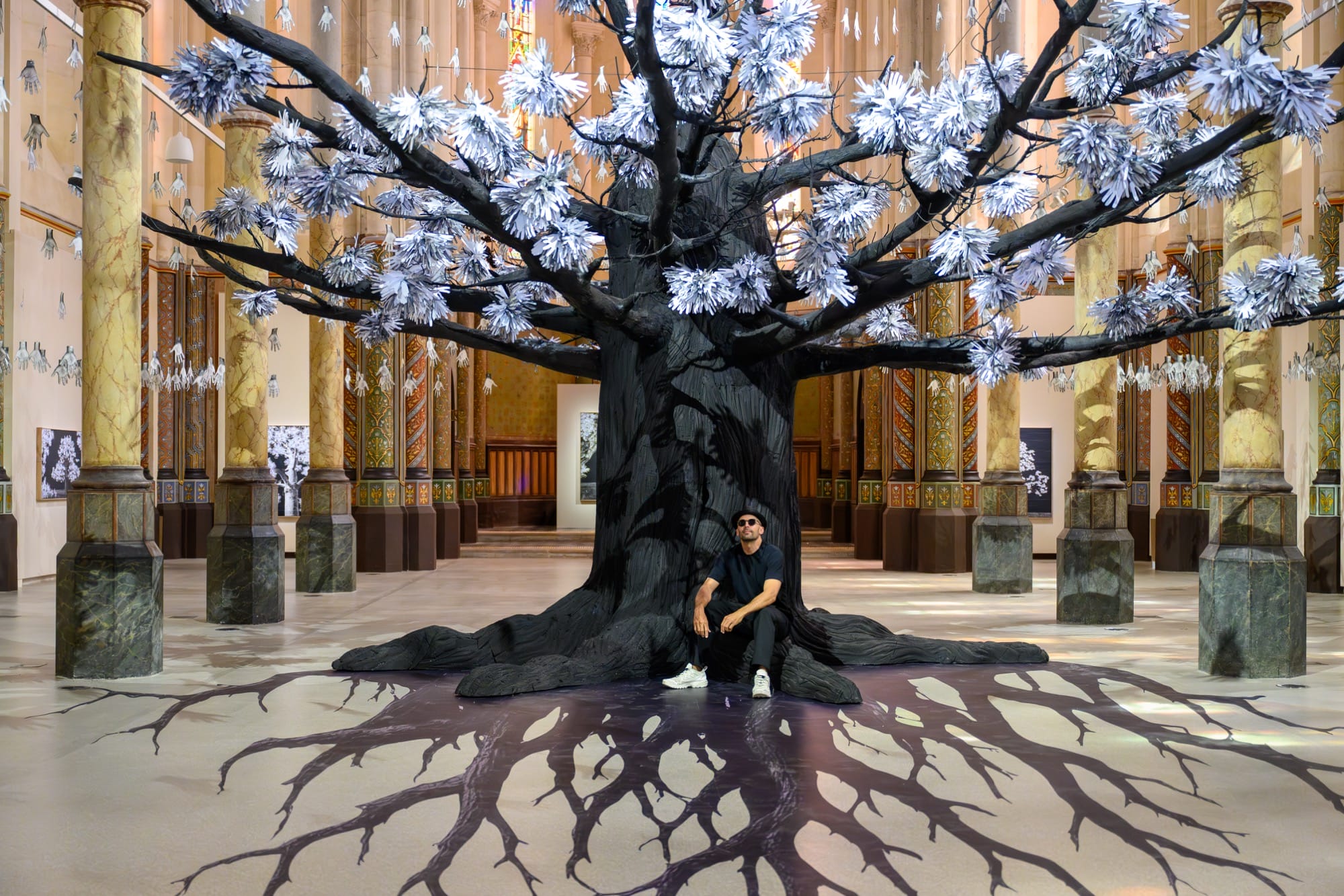 the artist sits underneath a tree installation with leaves made of hands