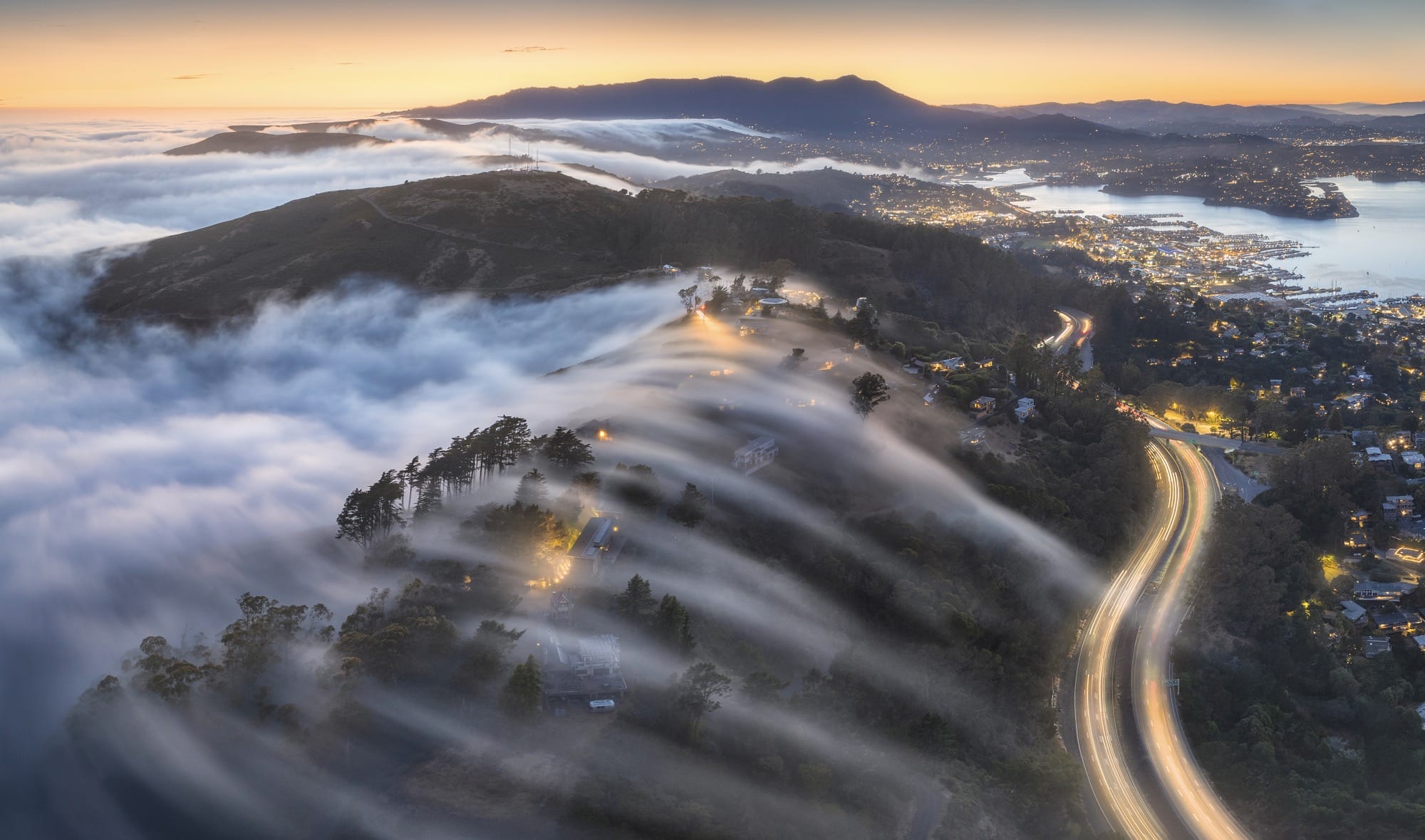 As the sun sets over the hills, soft waves of coastal fog drift like silk across the ridgeline, wrapping homes and trees in light. A glowing highway carves through the landscape like a lifeline.