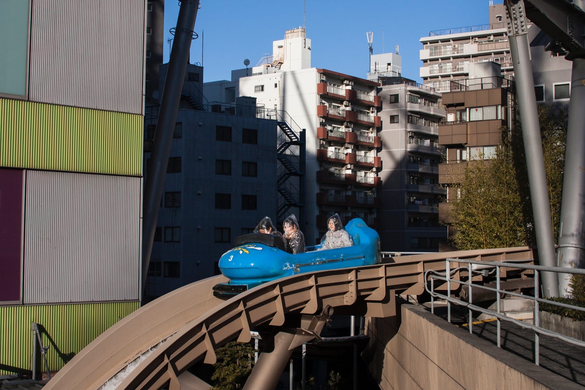 people ride on a blue rollercoaster car in a city, with their faces covered in plastic