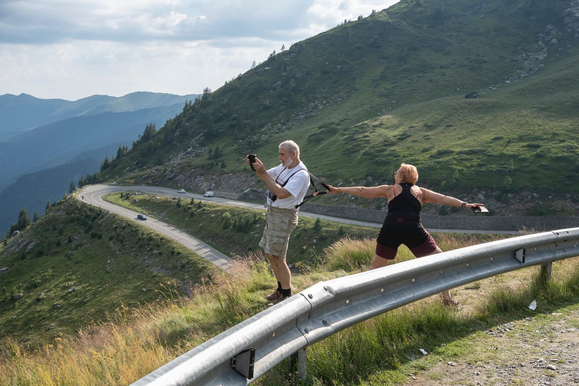 a couple stands on a mountain overlook, and the woman holds the man by his suspenders in case he slips