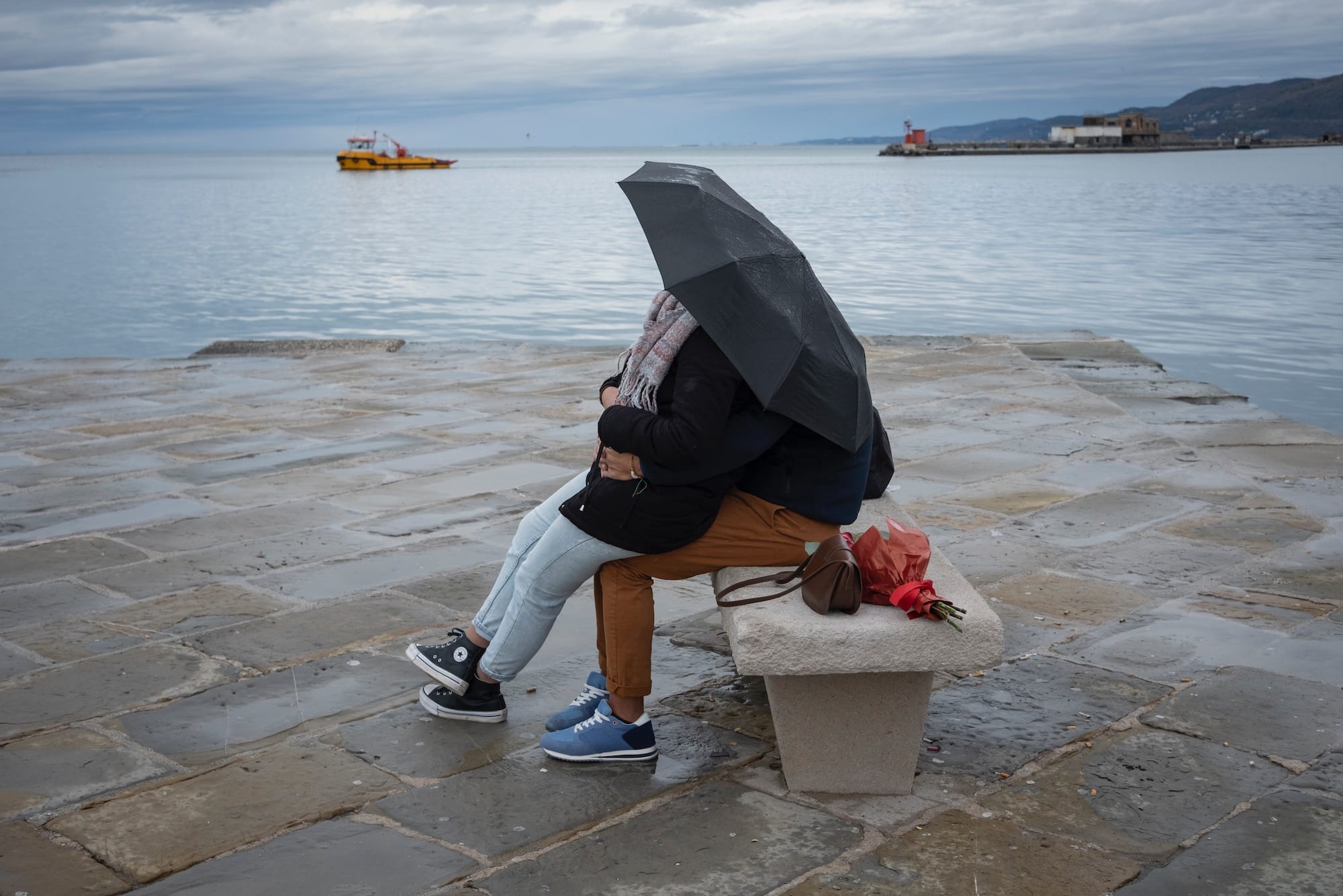 two people sit on a bench under an umbrella by the sea