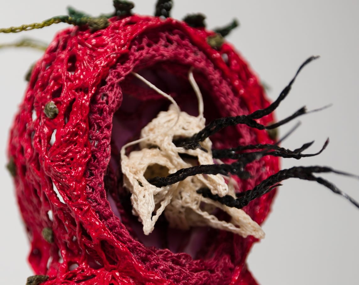 a detail of crocheted sculpture of skeletal baby bird heads with black strands coming out of their eyes, surrounded by a red nest