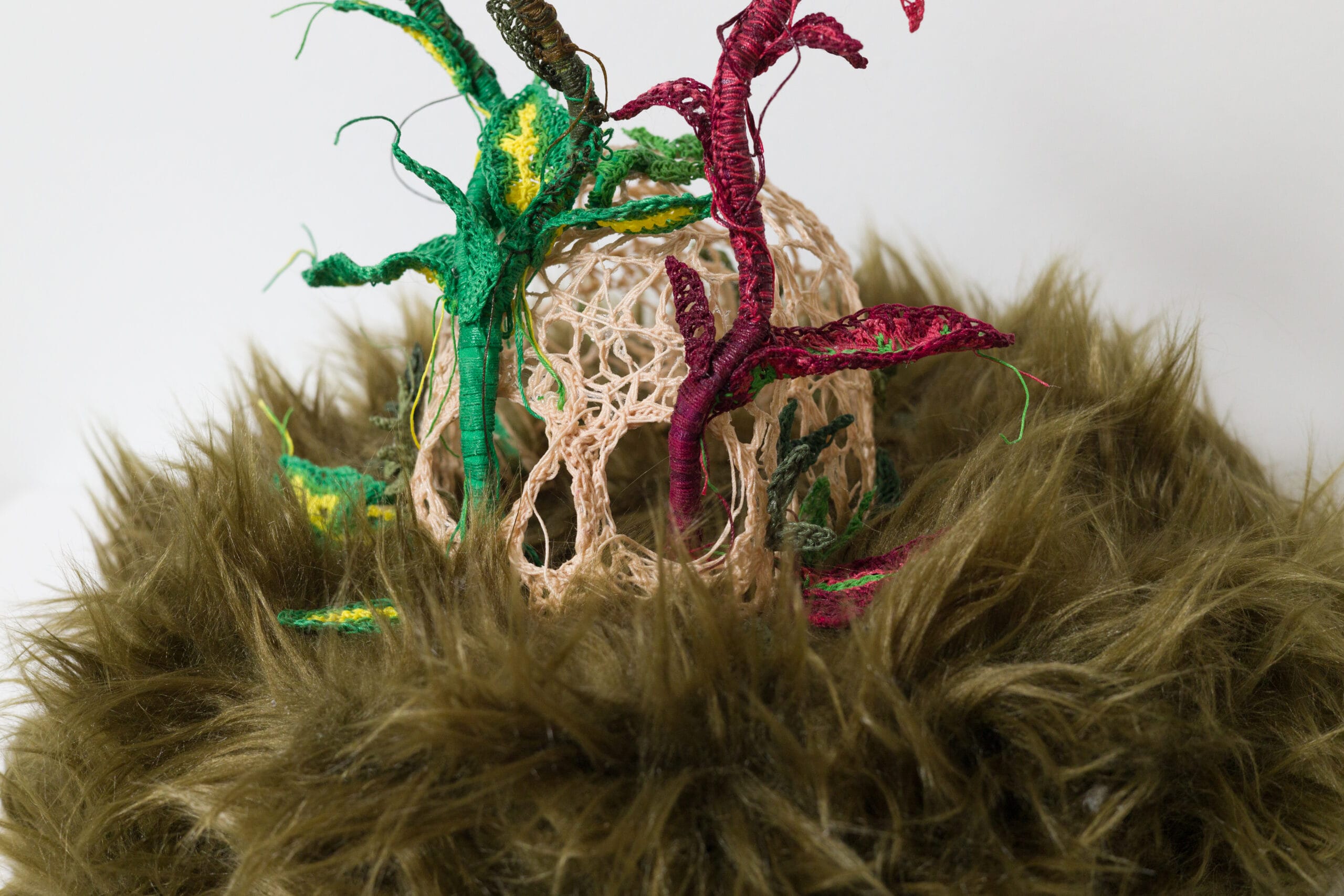 a detail of a textile sculpture featuring a crocheted human skill with plants growing out of the eyes