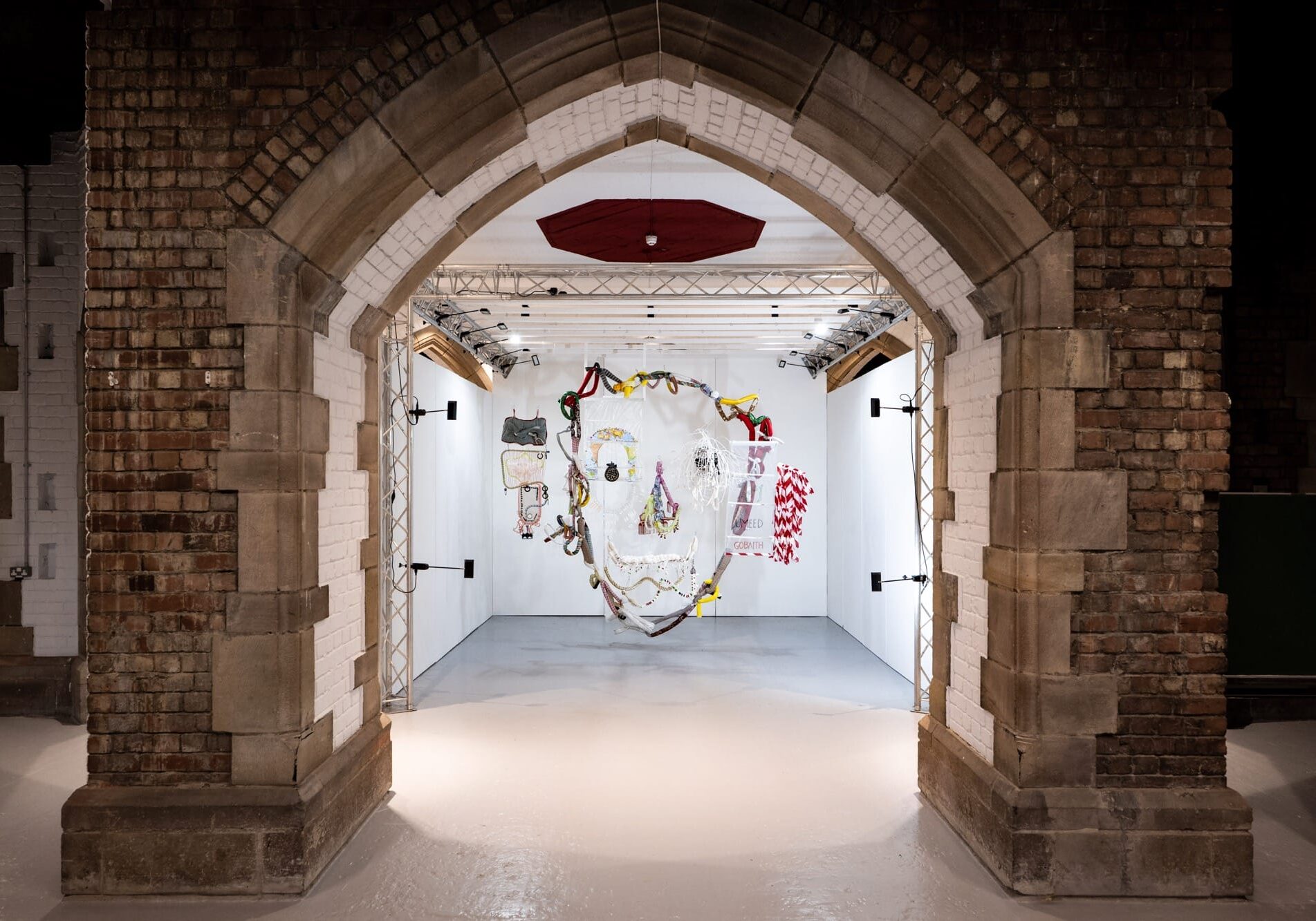 an installation view of an artwork made of various pieces of fabric and found objects, suspended from the ceiling to create a face when viewed straight-on