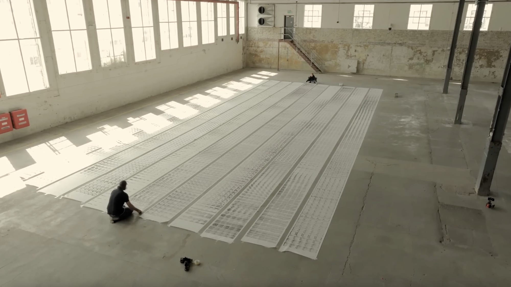 people stand around scrolls of patterned paper laying in rows on a concrete floor in a warehouse-type building