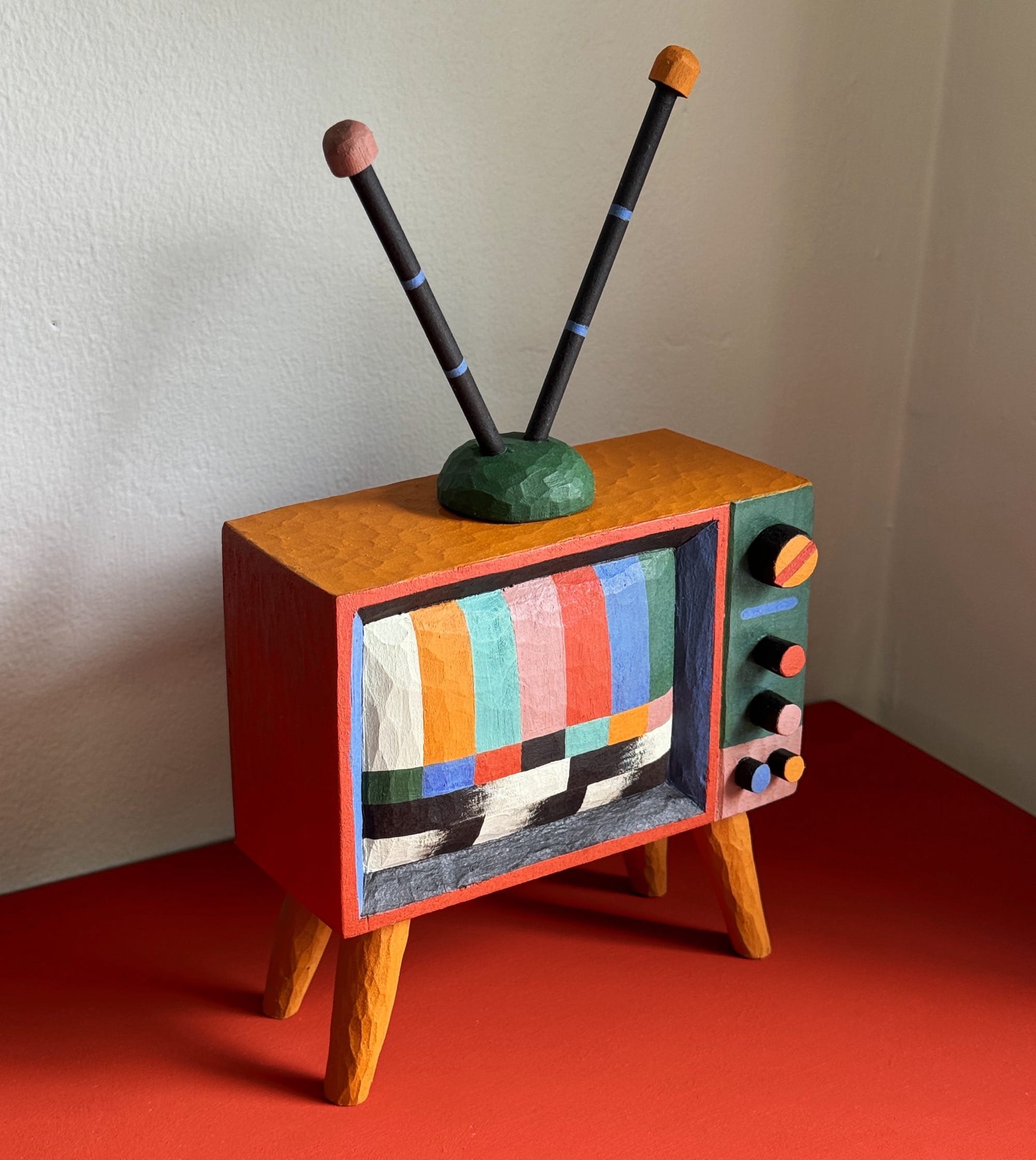 a colorful, hand-carved wooden sculpture of a retro TV with antennae and legs, with a test pattern on the screen